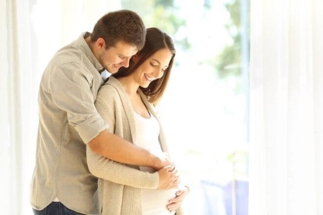 Schwangere Frau lehnt sich an Mann an. Mann legt Arme auf den Bauch der Frau. Beide schauen auf den Bauch.